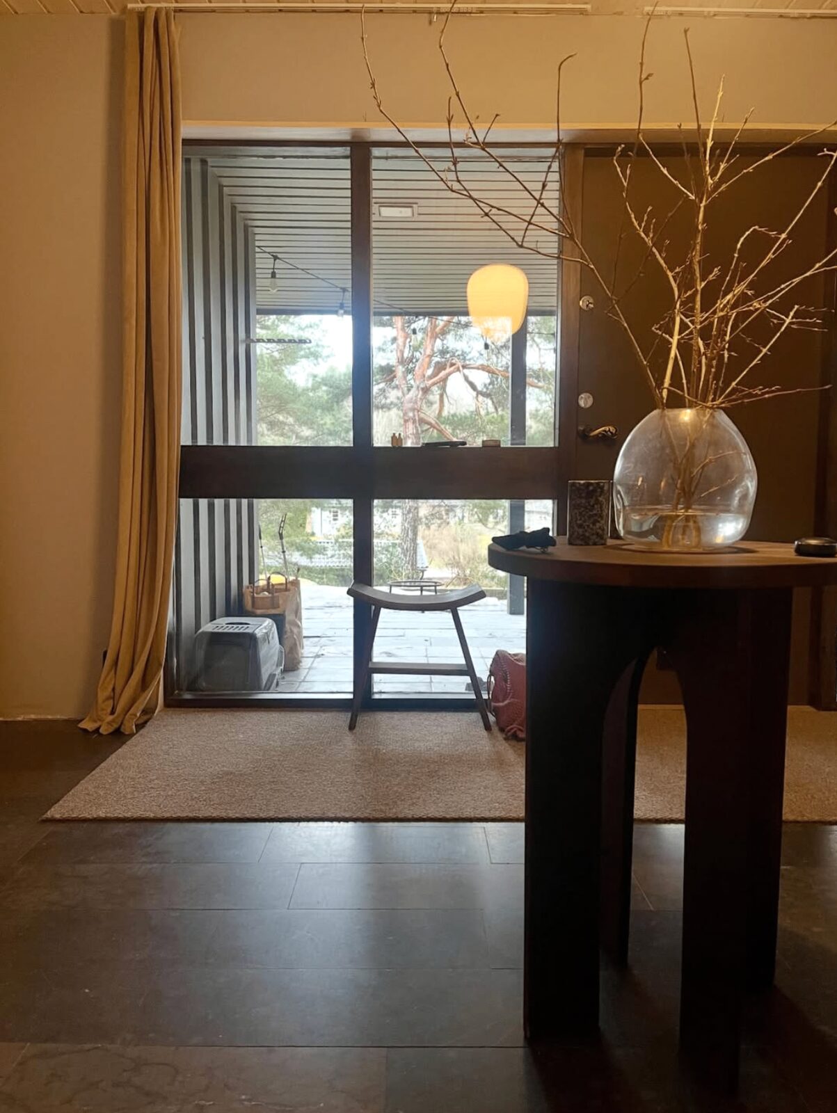 Runt sidobord i mörkt trä med valvformade ben och glasvas, placerat i en hall med solljus